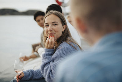 Smiling young woman with hand on chin sitting with friends on pier near lake