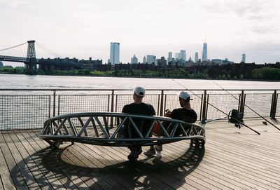 Rear view of men sitting on railing by river against sky