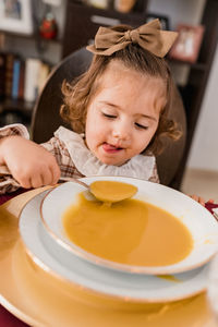 Charming child with bow on brown hair and spoon against plate of squash puree soup in house