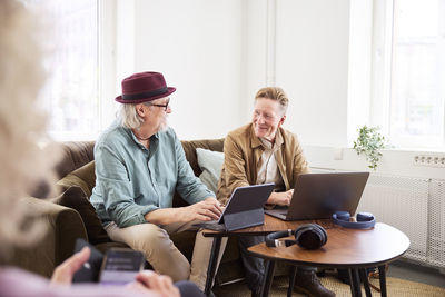 Senior men talking to each other while sitting in living room and using digital tablet and laptop to edit podcast