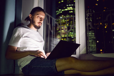 Freelance man in glasses working, sits at the window on the floor in front of a skyscraper at night