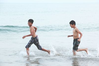 Side view of playful shirtless brothers running in sea on shore at beach