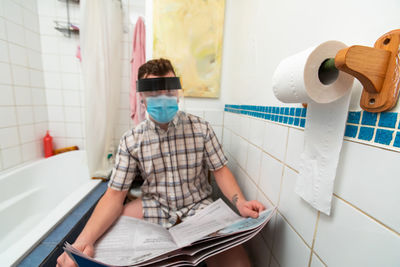 Man wearing mask reading newspaper while sitting in bathroom