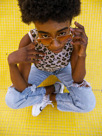 From above young african american female in trendy ripped jeans and stylish sunglasses sitting on yellow floor