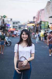 Portrait of smiling young woman with purse standing on street in city