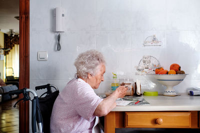 Side view of aged disabled female in wheelchair eating soup while sitting at table with food in light kitchen at home