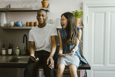 Cheerful young friends sitting with smart phones on kitchen counter at home