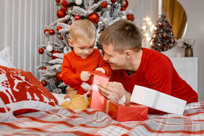 Happy smiling father and his little son unwrapping a present sitting on the bed near christmas tree