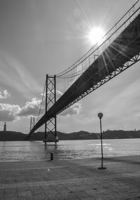 Low angle view of april 25th bridge over river against cloudy sky on sunny day