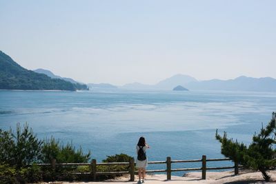 Full length rear view of woman standing by river against clear sky on sunny day