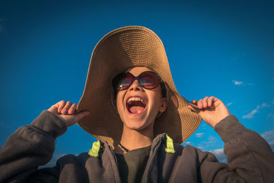 Low angle view of cheerful boy wearing hat while standing against blue sky