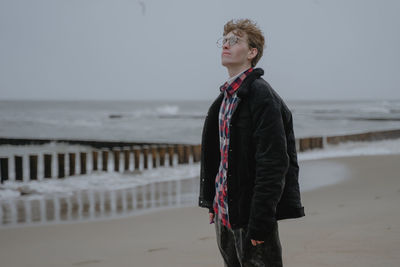 A young man with glasses is standing, enjoying the sea air, looking up, in a red check shirt