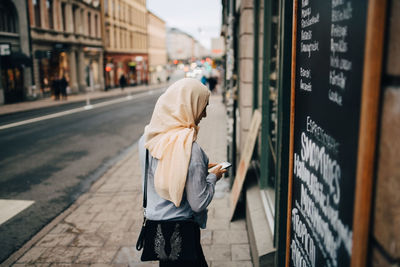 Young woman using smart phone while entering cafe in city