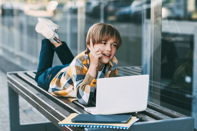 Boy using laptop while lying on bench by modern building