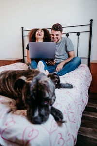 Young couple using laptop while dog sleeping on bed at home