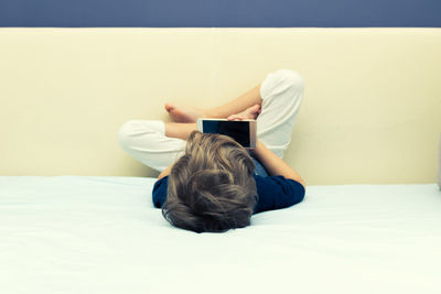 Rear view of boy using mobile phone on bed against wall