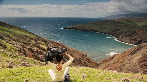 Woman looking at sea while sitting on mountain against sky