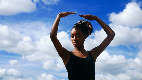 Portrait of young woman with arms raised standing against sky