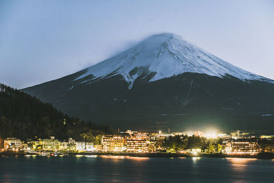 Illuminated buildings by snowcapped mountain against sky at night