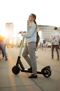Young woman looking away while standing on push scooter against sky in city
