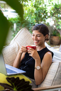 Happy young woman working remotely on laptop and making video call, drinking cappuccino