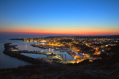 High angle view of illuminated city by sea against sky at sunset