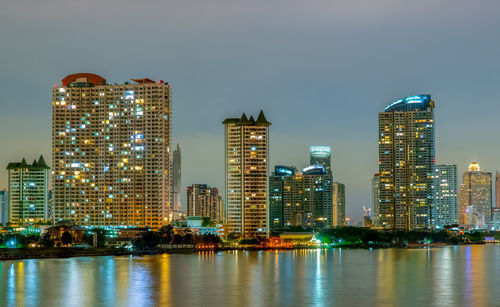 Illuminated buildings by river in city against sky at night