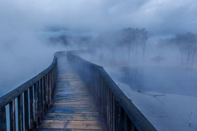 Wooden footbridge over frozen lake against sky during foggy weather