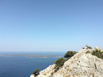 Rear view of people standing on rock by sea against clear blue sky