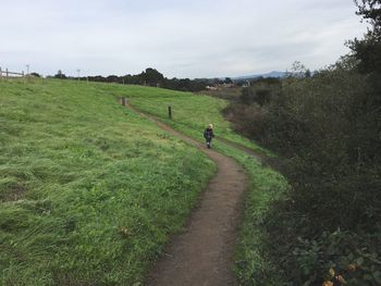 Mid distance view of boy walking on trail amidst field against cloudy sky
