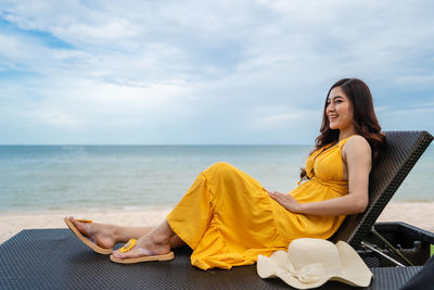Smiling woman looking away while sitting against sky at beach