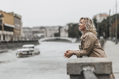 A middle-aged blonde woman with curly hair stands on the river embankment in the city.