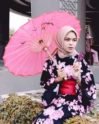 Woman holding pink umbrella and hand fan while sitting outdoors