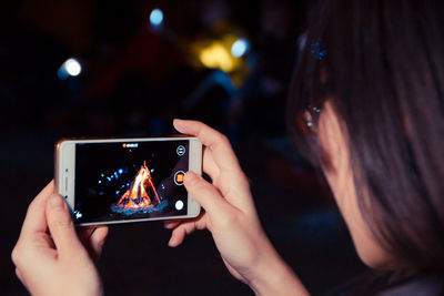 Cropped image of woman filming bonfire through mobile phone at night