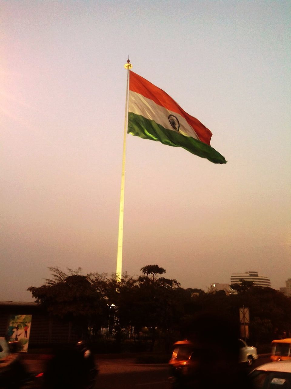 transportation, clear sky, flag, sunset, sky, mode of transport, built structure, city, incidental people, car, architecture, building exterior, outdoors, low angle view, flying, travel, tree, dusk, patriotism, national flag