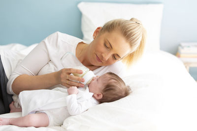Mother feeding milk with bottle to baby girl on bed at home