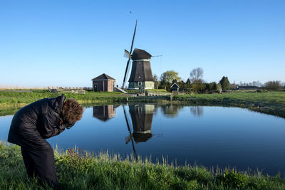 Person photographing by lake against windmill and blue sky