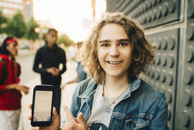 Portrait of smiling young man showing mobile phone while friends standing in background on street
