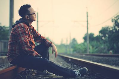 Side view of young man sitting on railroad track against sky