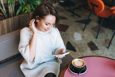 Attractive young brunette woman in white casual dress with cup of coffee using mobile phone in cafe