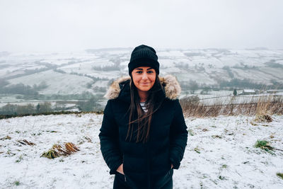 Portrait of young woman wearing knit hat while standing on snow field against sky