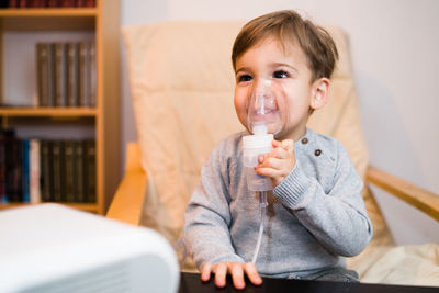 Smiling cute boy inhaling through respiratory mask while sitting on chair at home