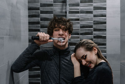 Portrait of man brushing teeth while standing with smiling woman in bathroom