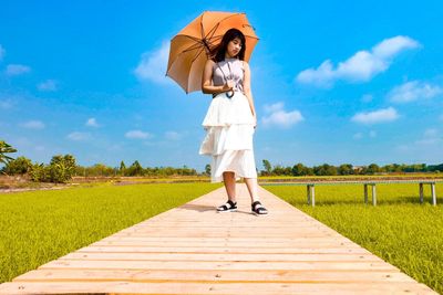Full length of woman holding umbrella while standing on boardwalk over field against sky
