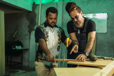 Concentrated multiethnic male colleagues in casual wear sawing wooden board at table while working together in light joinery with professional equipment