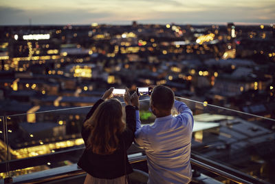 Rear view of male and female professionals photographing city on mobile phone from terrace during sunset