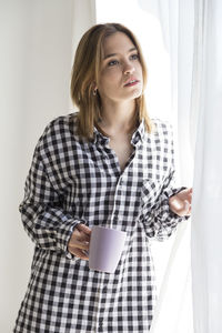Young woman holding coffee cup by curtains at home
