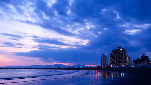 Illuminated buildings by sea against sky at dusk