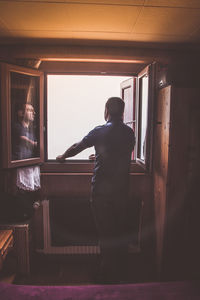 Rear view of young man standing against window at home