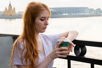 Pensive young woman holding paper cup with coffee while standing near river